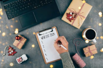 Woman's hand writing 2024 Wish list in notebook. Laptop, present box, cup of coffee